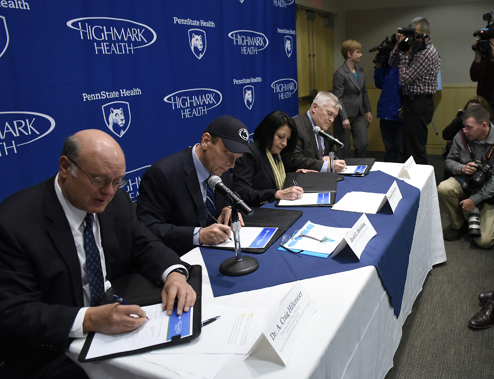 Four people - three men and a woman - wearing business attire sit at a table, each signing a document. A background with the Penn State Health and Highmark Health logos is behind them.