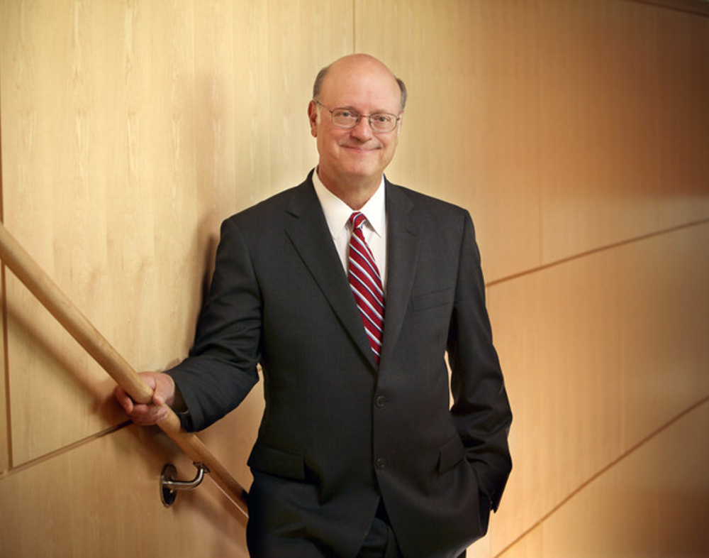 Dr. Craig Hillemeier wears a suit and poses against a wooden wall. His right hand is on the banister, his left hand is in his pocket.
