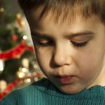 A close-up of a young boy in a green sweater, looking downward. A Christmas tree is in the background, slightly out of focus.