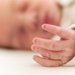 A close-up of an infant™s hand. The baby™s face is seen in the background, out of focus. The baby is sleeping.
