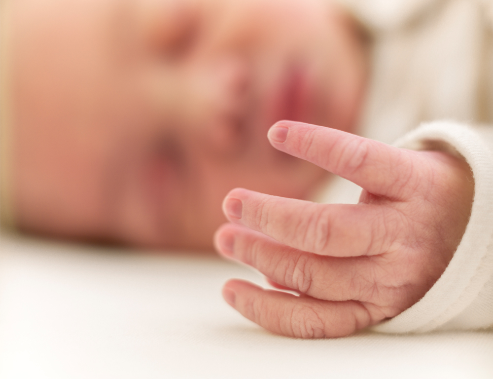 A close-up of an infant™s hand. The baby™s face is seen in the background, out of focus. The baby is sleeping.