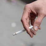A close-up of a hand holding a lit cigarette. The background is out of focus.