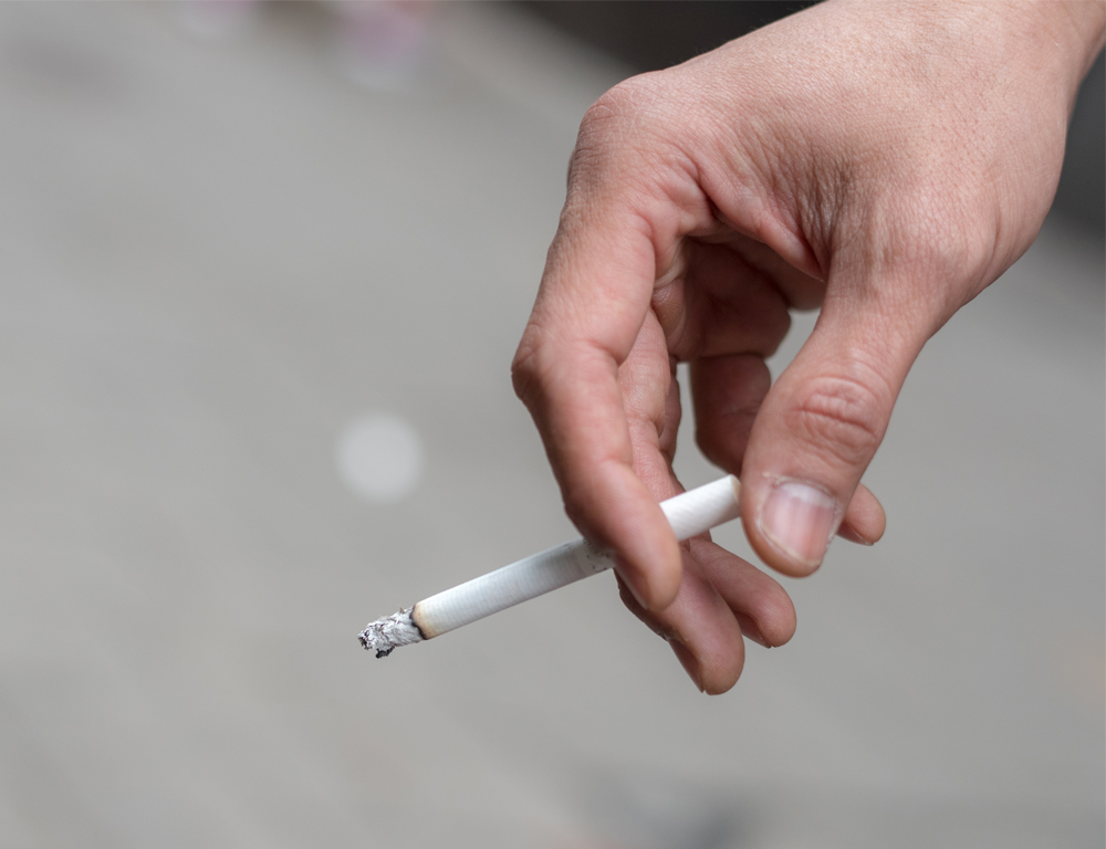 A close-up of a hand holding a lit cigarette. The background is out of focus.