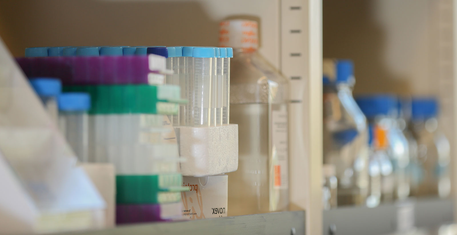 A close-up image of laboratory equipment depicts test tubes, boxes and bottles on the shelf in a Penn State College of Medicine laboratory in summer 2016. Test tubes and one bottle are visible at left in focus, with other bottles out-of-focus to the right.