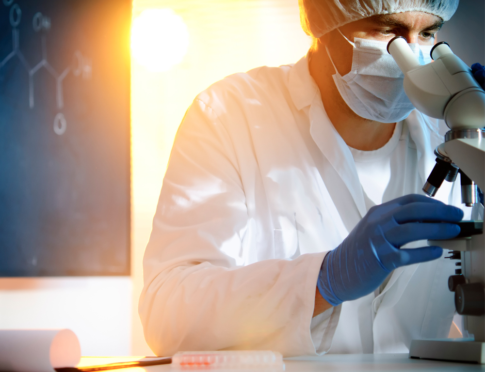 A lab technician in a white coat, blue gloves and a mask and cap looks into a microscope, most of which sits out of the photo to the right. A bright light shines over the individual's shoulder.