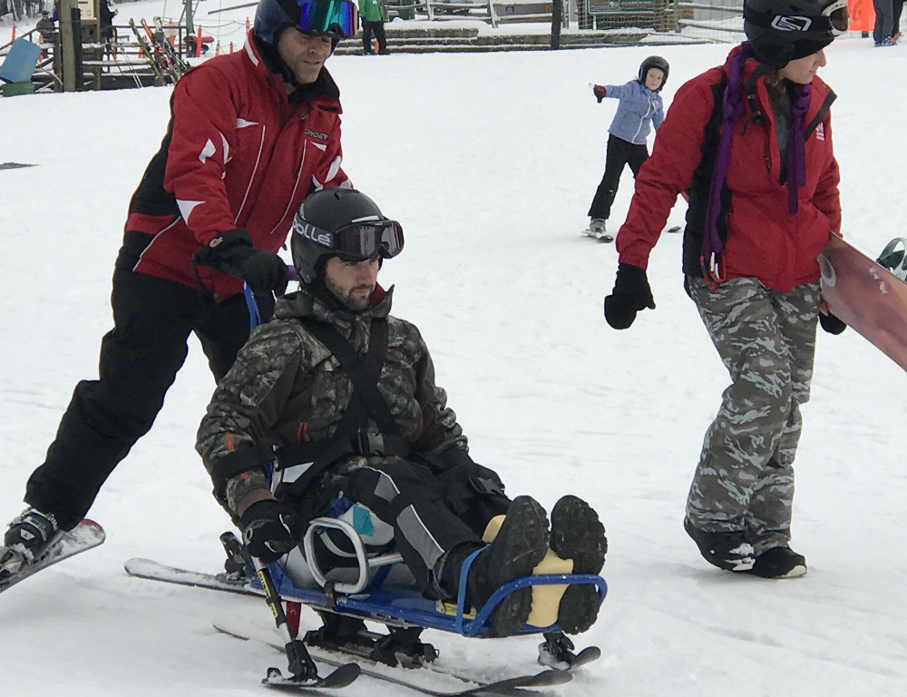 Two people in winter coats and ski pants help guide a man who's sitting on a sled move across a ski slope.