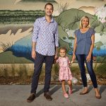 Erin Weidemann, right, is seen with her husband, Brent Weidemann, and their daughter, Rooney Cruz Weidemann. The three are pictured standing in front of a wall with a mural painted on it, and are holding hands.