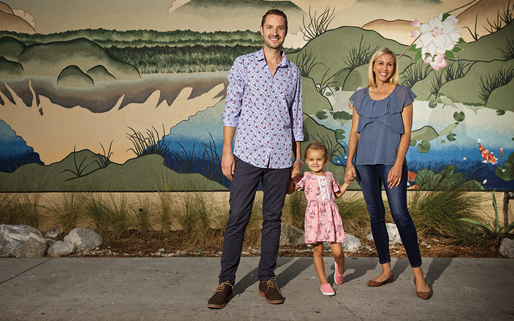 Erin Weidemann, right, is seen with her husband, Brent Weidemann, and their daughter, Rooney Cruz Weidemann. The three are pictured standing in front of a wall with a mural painted on it, and are holding hands.