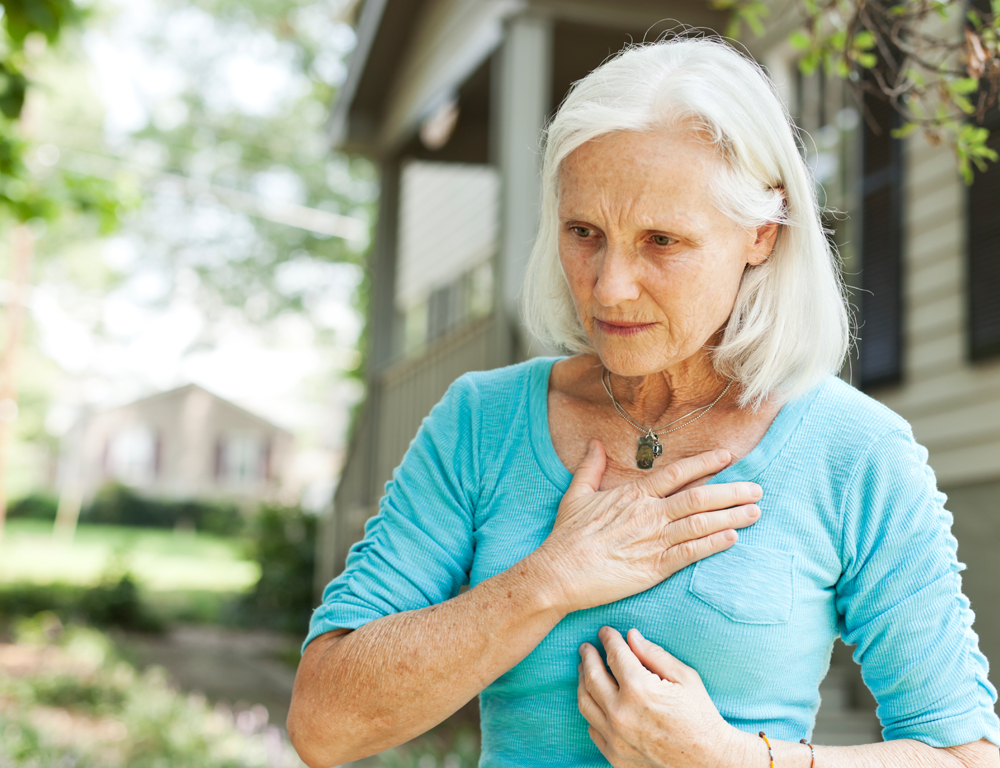 A woman in a blue shirt stands outside of a home, which is slightly out of focus in the background. The woman holds her hand to her chest, up near her neck.