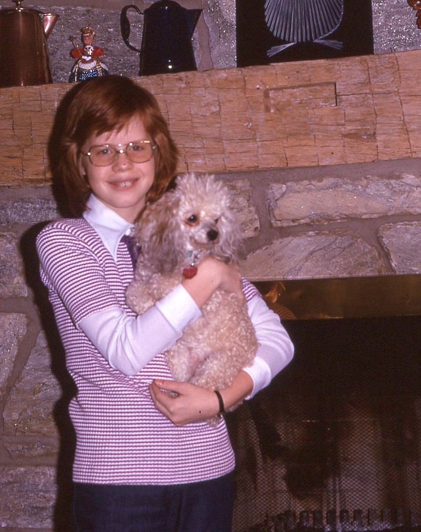 Girl with brown hair and glasses holds poodle while standing by fireplace.