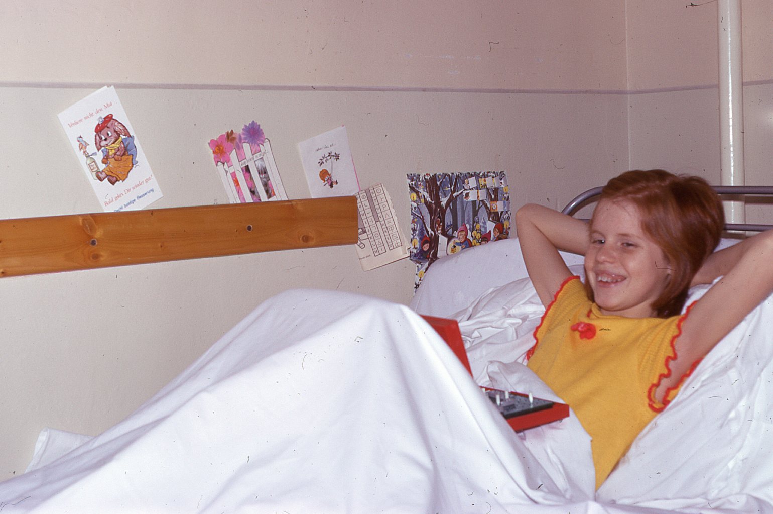 Girl with red hair smiles while lying in hospital bed and holding a game on her lap.