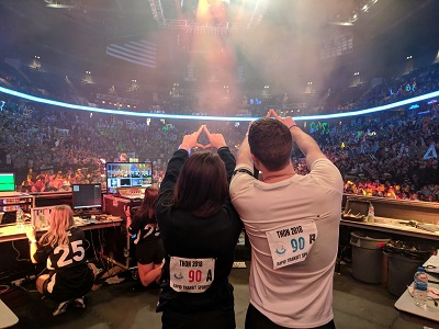 Two students, one female and one male, stand with their backs to the camera, facing an arena full of people. They stand with their hands in the air, making a diamond shape with their fingers.