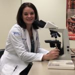 Katie Schieffer, a woman with shoulder-length brown hair wearing a white lab coat is smiling while seated at a table. Her hands are on either side of a microscope with a case of slides sitting next to the microscope. A poster of the digestive system is on the wall behind her.