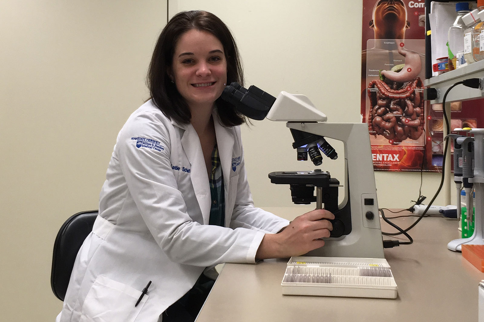 Katie Schieffer, a woman with shoulder-length brown hair wearing a white lab coat is smiling while seated at a table. Her hands are on either side of a microscope with a case of slides sitting next to the microscope. A poster of the digestive system is on the wall behind her.