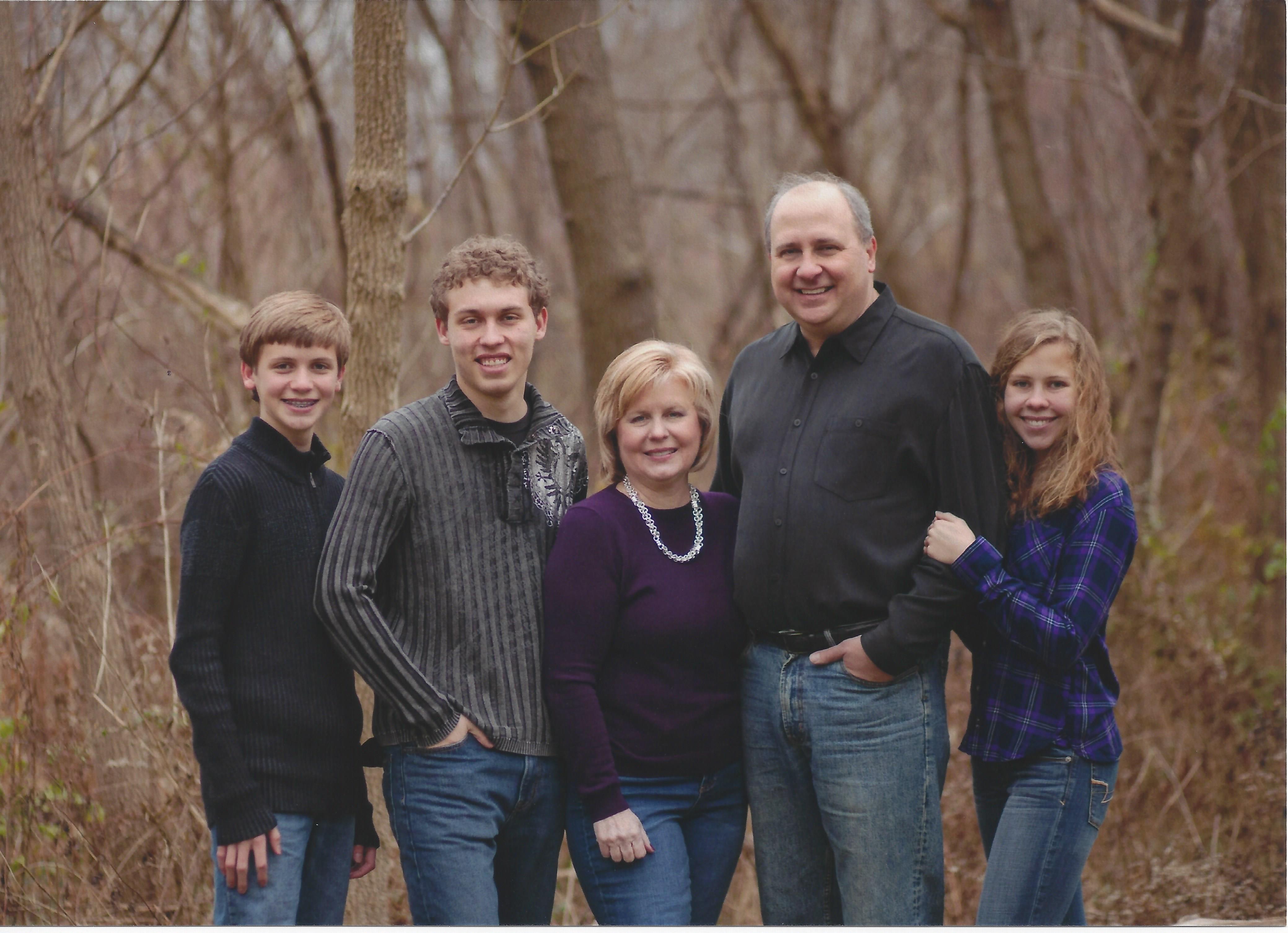 Family of five pose outdoors in front of a wooded scene. Family includes two boys, a woman in a purple top, a man in a gray shirt and a girl.