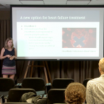 Scott Graupensperger and Bethany Latten give a presentation during the 2017 summer Translational Science Fellowship, a highly interactive program for both medical and graduate students to receive a research foundation. They are pictured standing, facing a row of chairs, with the audience visible from behind. A presentation screen is to their left, with a slide titled "A new option for heart failure treatment" visible.