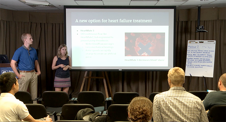 Scott Graupensperger and Bethany Latten give a presentation during the 2017 summer Translational Science Fellowship, a highly interactive program for both medical and graduate students to receive a research foundation. They are pictured standing, facing a row of chairs, with the audience visible from behind. A presentation screen is to their left, with a slide titled 