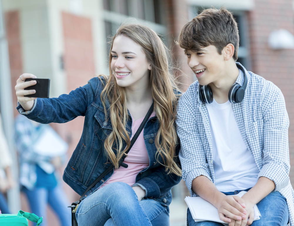 A teenage boy and girl sit together and look into a phone the girl is holding up, apparently posing for a selfie. A brick building with windows is in the background.