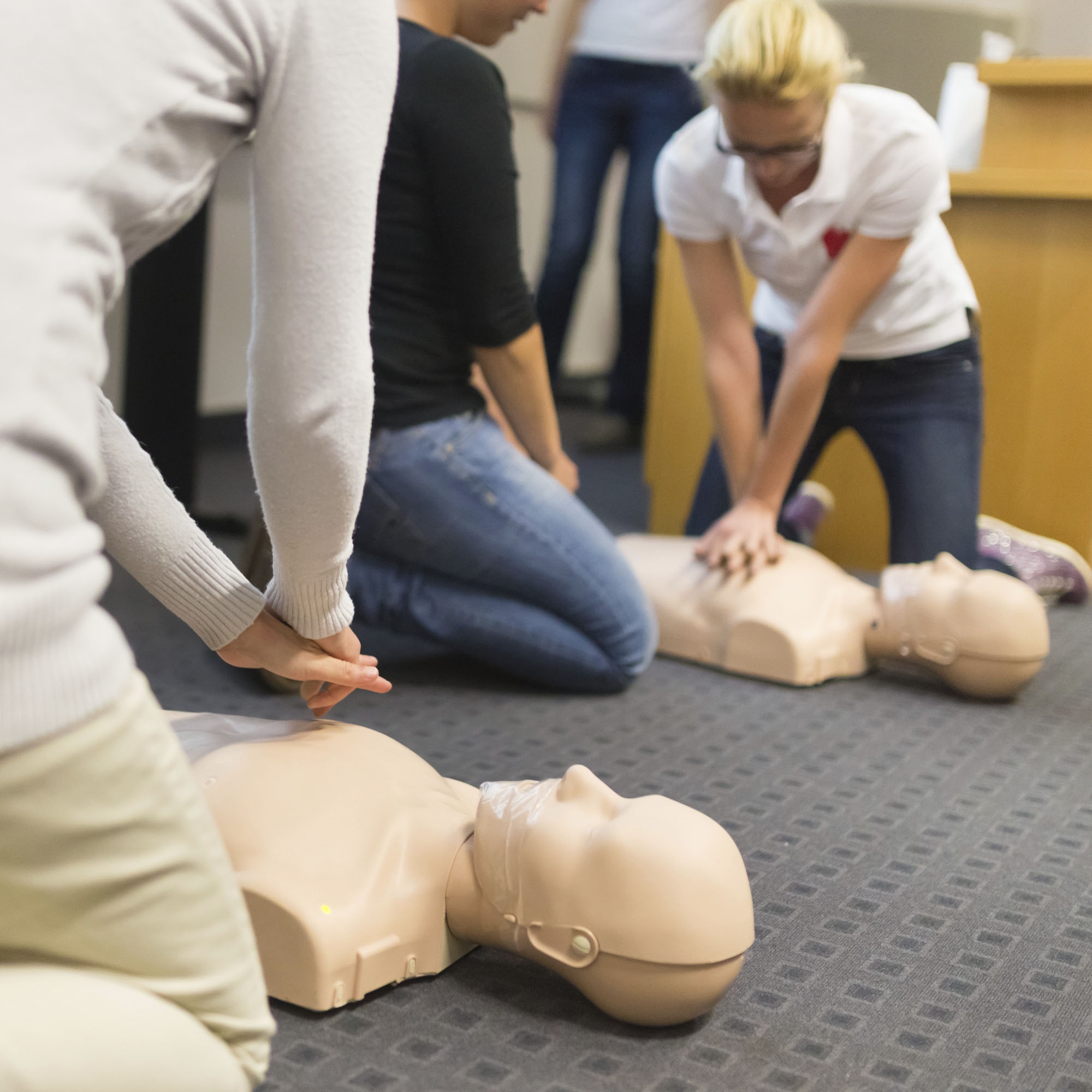 Two women practice CPR chest compressions on dummies while one person watches. The dummies lie on a carpet with a square-checked pattern. Another person stands in the background.