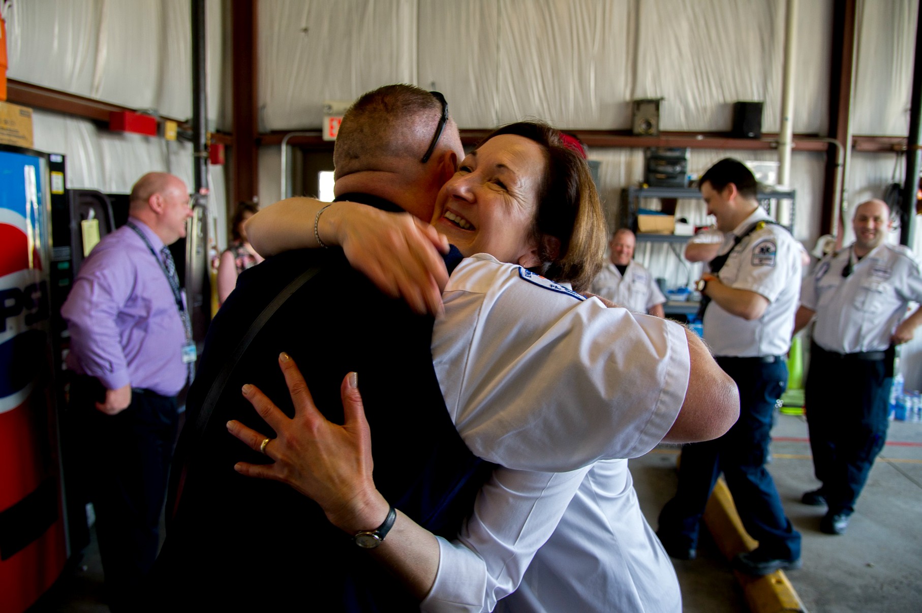 Cardiac arrest survivor Marty Woodfin smiles as she hugs Life Lion Paramedic Jeff Gewertz in the Life Lion garage. In the background, other paramedics smile and talk.