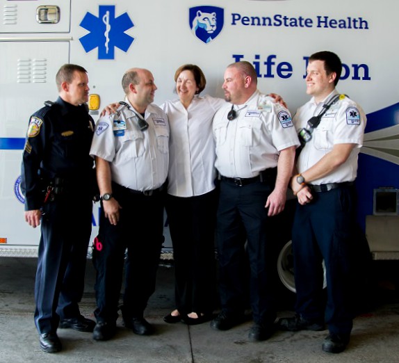 Marty Woodfin is seen standing between EMT Robert Snyder and Paramedic Jeff Gewertz, with an arm around each. She is smiling and looking at Snyder. Derry Township Police Sgt. Eric Singiser and EMT Justin McNaughton are looking at Woodfin and smiling. They are standing in front of a Life Lion ambulance.
