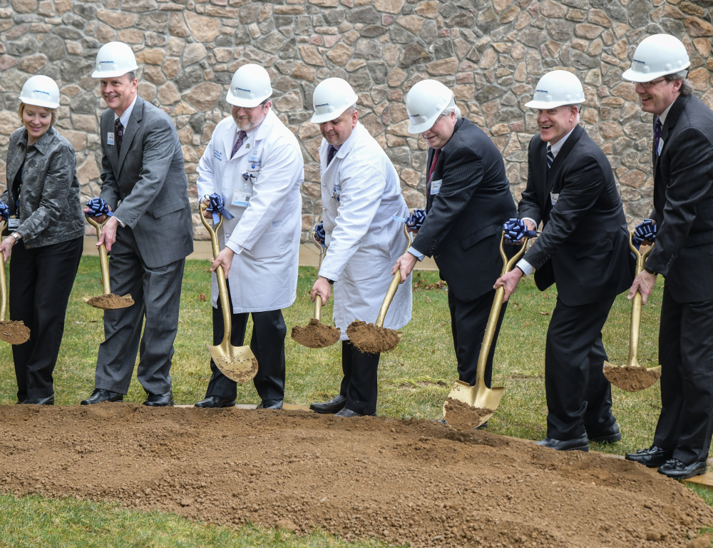 Seven people -- five in business suits, two in white physician's coats, and all wearing white helmets -- use gold shovels to dig into a pile of dirt. A stone wall is in the background.