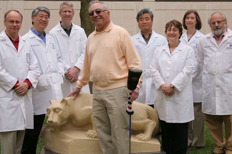 Warren Gittlen, center, holding golf club, stands in front of a statue of the Nittany Lion with seven Penn State researchers. They are outdoors. The researchers, a mix of both men and women, are wearing white lab coats.