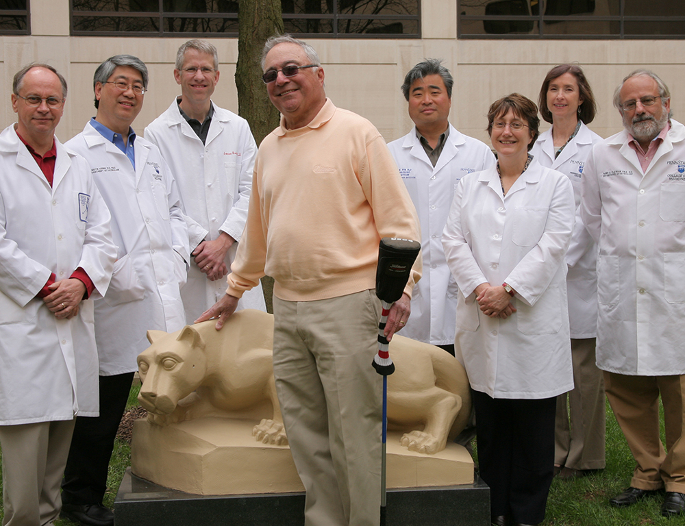 Warren Gittlen, center, holding golf club, stands in front of a statue of the Nittany Lion with seven Penn State researchers. They are outdoors. The researchers, a mix of both men and women, are wearing white lab coats.