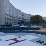 The Life Lion helipad at Hershey Medical Center is in the foreground, with a large white cross with a red "H" in the middle. The Medical Center's signature Crescent building is in the background.