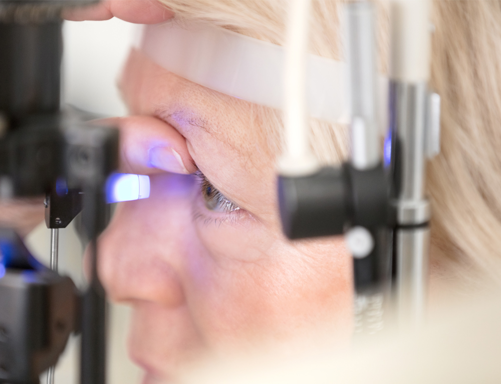 A close-up of a woman with her head placed against a piece of eye exam equipment. The equipment is shining a bright light into her eye, which is being held open by her thumb.