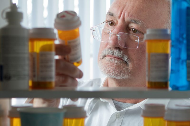 View from inside a medicine cabinet looking out, as a man in a white shirt holds an orange-brown pill bottle and looks at it. Other similar bottles are in the foreground.