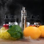 A vaping device sits on a table, surrounded by several dropper bottles with various colors of fluid. In the foreground are grapes, lemons and a green leaf.