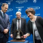 Rosemary Manbachi, a Penn State Cancer Institute patient, rings a bell symbolizing the end of an individual's cancer treatment. Looking on are David L. Holmberg, president and CEO of Highmark Health (left), and Dr. Ray Hohl, director of Penn State Cancer Institute (center). A blue backdrop contains the Highmark Health and Penn State Health logos.