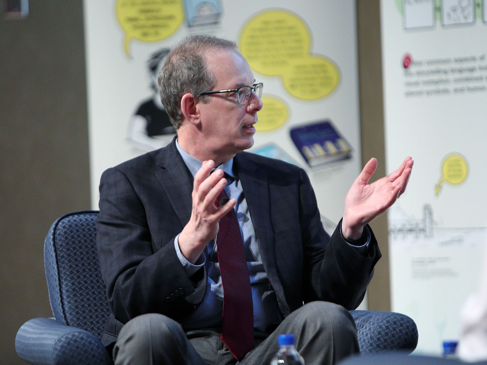 Dr. Michael Green recently discussed how reading and creating comics helps students with practical doctoring skills during a panel discussion at the National Institutes of Health. Green is pictured seated, gesturing with his hands and looking to the right. Large posters are visible behind him.