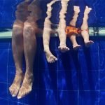 View from underwater of three pairs of legs. All three people are sitting on the edge of the pool. The side of the pool is blue tile.