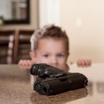 A handgun sits on a counter in the foreground. In the background, a child, slightly out of focus, peers over the counter at the gun.