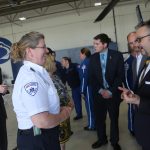 A woman in an EMS uniform speaks with a man in a suit. A helicopter is mostly off-camera, to the right. Other people are in the area “ a helicopter hangar “ standing and talking.