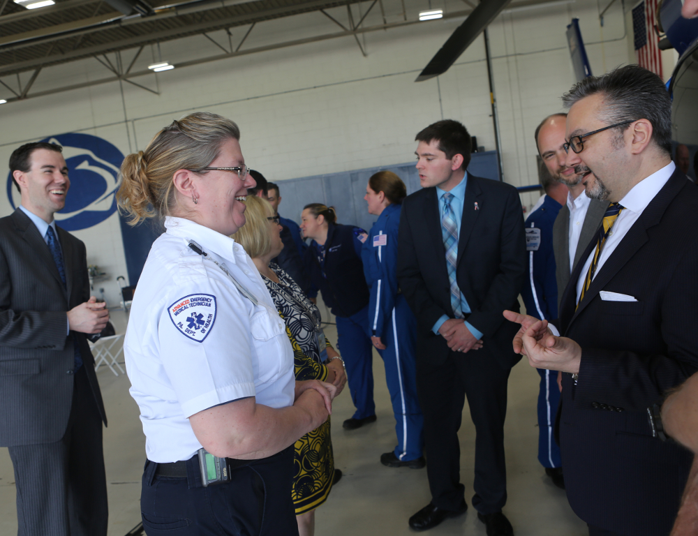 A woman in an EMS uniform speaks with a man in a suit. A helicopter is mostly off-camera, to the right. Other people are in the area “ a helicopter hangar “ standing and talking.