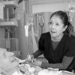 A female nurse leans over a male patient's bed while holding onto a ventilation tube. Various medical equipment is in the background.