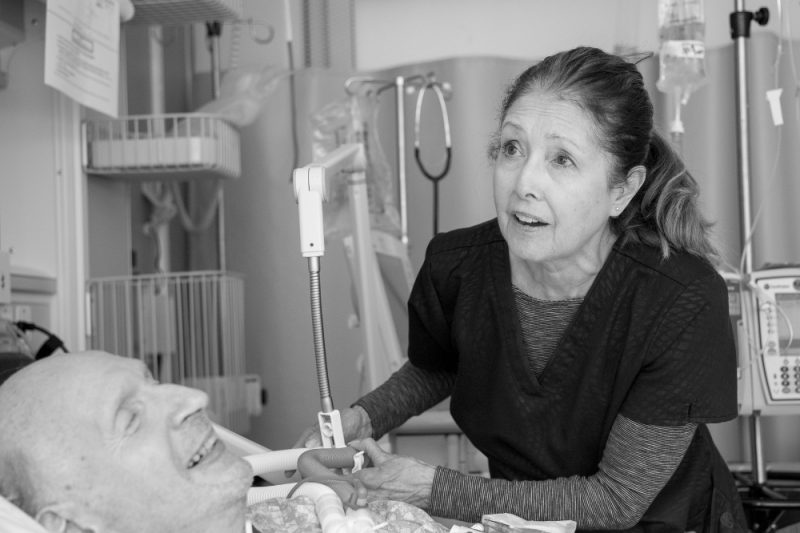 A female nurse leans over a male patient's bed while holding onto a ventilation tube. Various medical equipment is in the background.