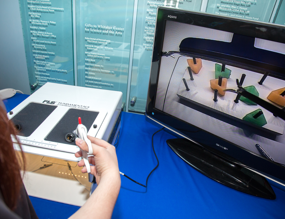 A close-up of a person™s right-hand holding a device that™s part of a table-top surgery simulator. A monitor is to the right of the device.