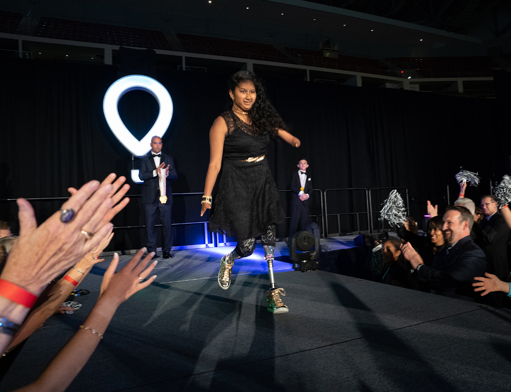 A young girl with a prosthetic leg walks on a stage as people standing around the stage applaud. The CMN (balloon) logo is in the background.