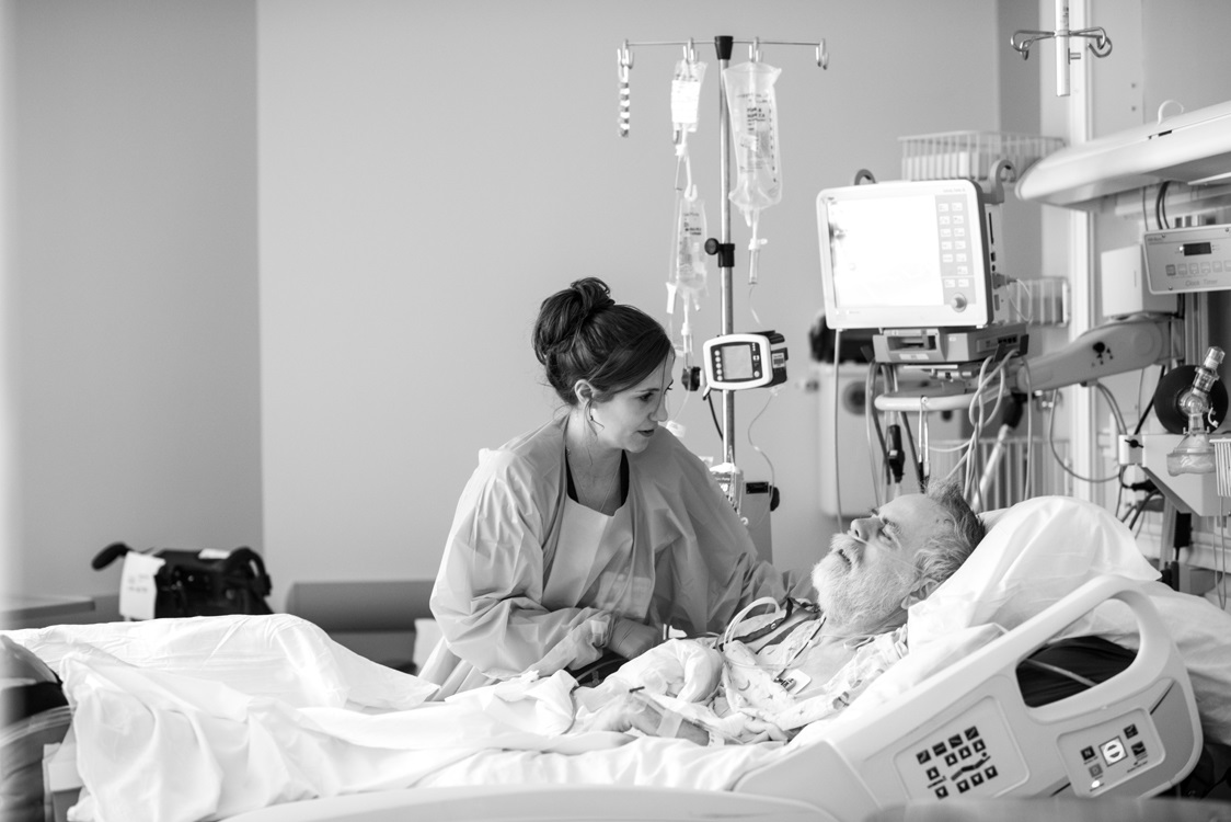 Mackenzie Bosse, a nurse in the Medical Intensive Care Unit at Penn State Health Milton S. Hershey Medical Center, leans over an older male patient and holds his hand. He is lying in a hospital bed and has a nasal cannula device for oxygen in his nose. A saline bag and other medications hang from a portable IV stand behind them, and a vital signs monitor is above the patient.