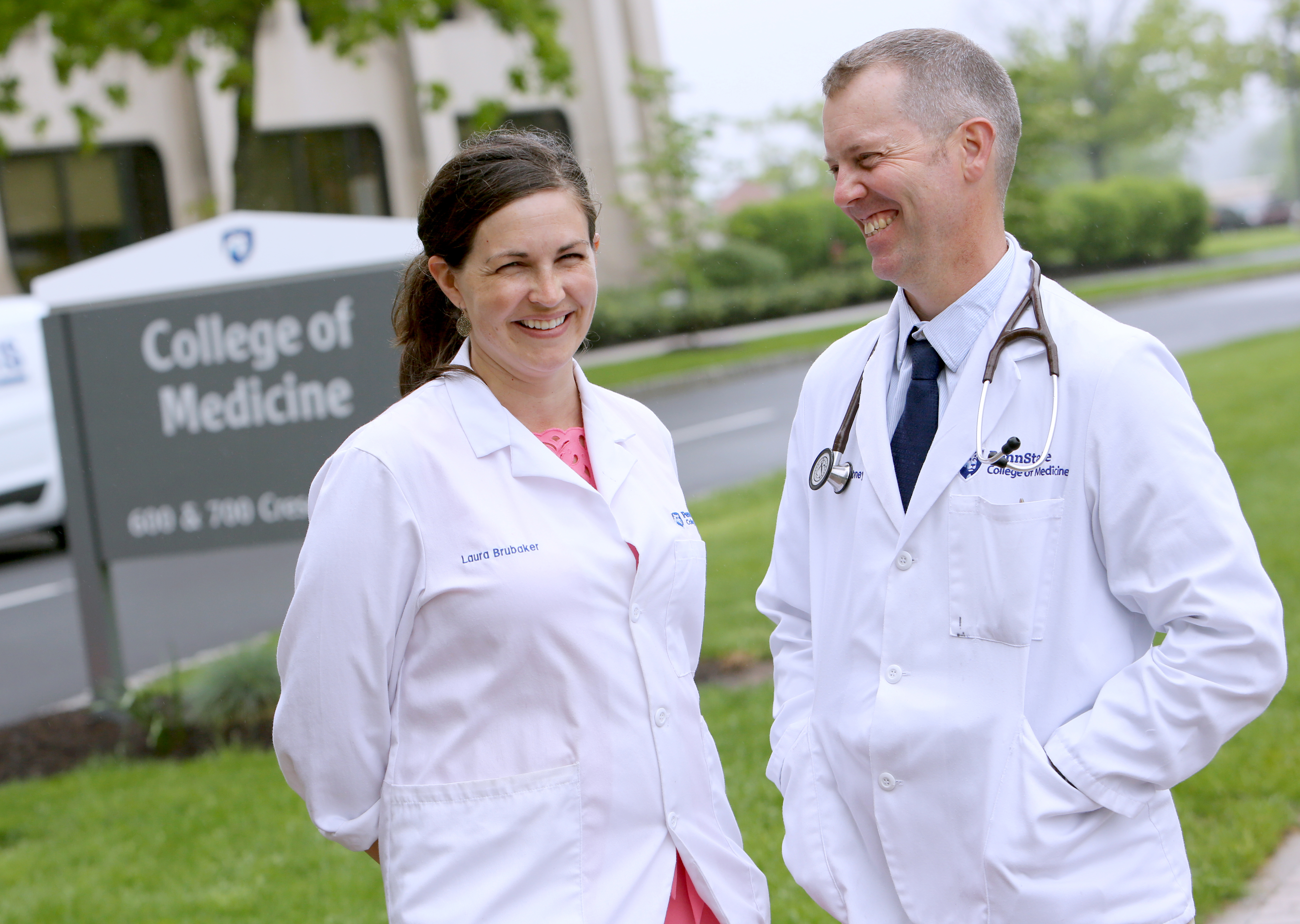 Dr. Laura Brubaker, left, and Dr. Benjamin Abney smile in front of the Penn State College of Medicine sign. They are wearing physician lab coats with their names and the College of Medicine logo on them. Dr. Abney has a stethoscope around his neck. Behind them is a road and the College of Medicine building.