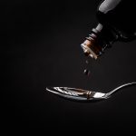A close-up of liquid being poured out of a dark bottle onto a spoon, against a black backdrop.