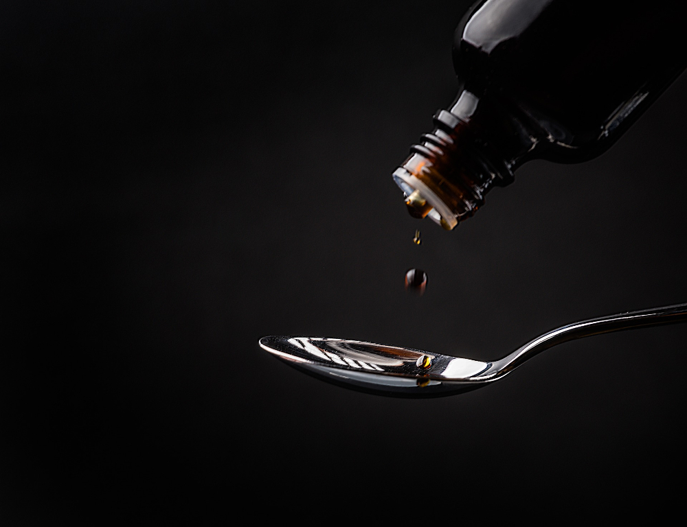 A close-up of liquid being poured out of a dark bottle onto a spoon, against a black backdrop.