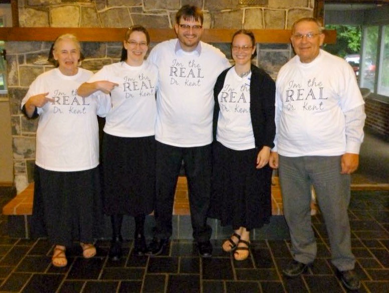 Dr. James Kent in the middle, his sisters next to him, his mother on the outer left, and his father on his outer right, pose in front of a fieldstone wall, all wearing identical white t-shirts with blue text that proclaims, “I’m the Real Dr, Kent.” All five family members are physicians and Penn State College of Medicine graduates.