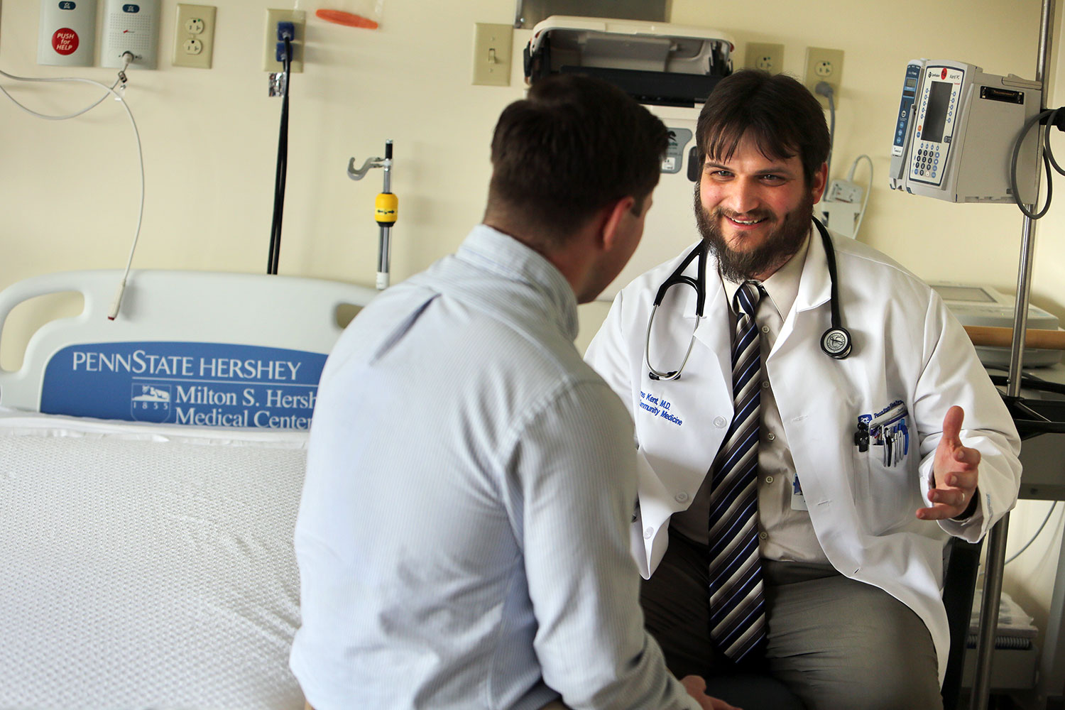 Dr. James Kent, a medium-height white male with brown hair and a beard dressed in a white lab coat and wearing a stethoscope around his collar, counsels a male patient seated on the edge of his hospital bed. The male patient has short, dark hair and is wearing an Oxford shirt.