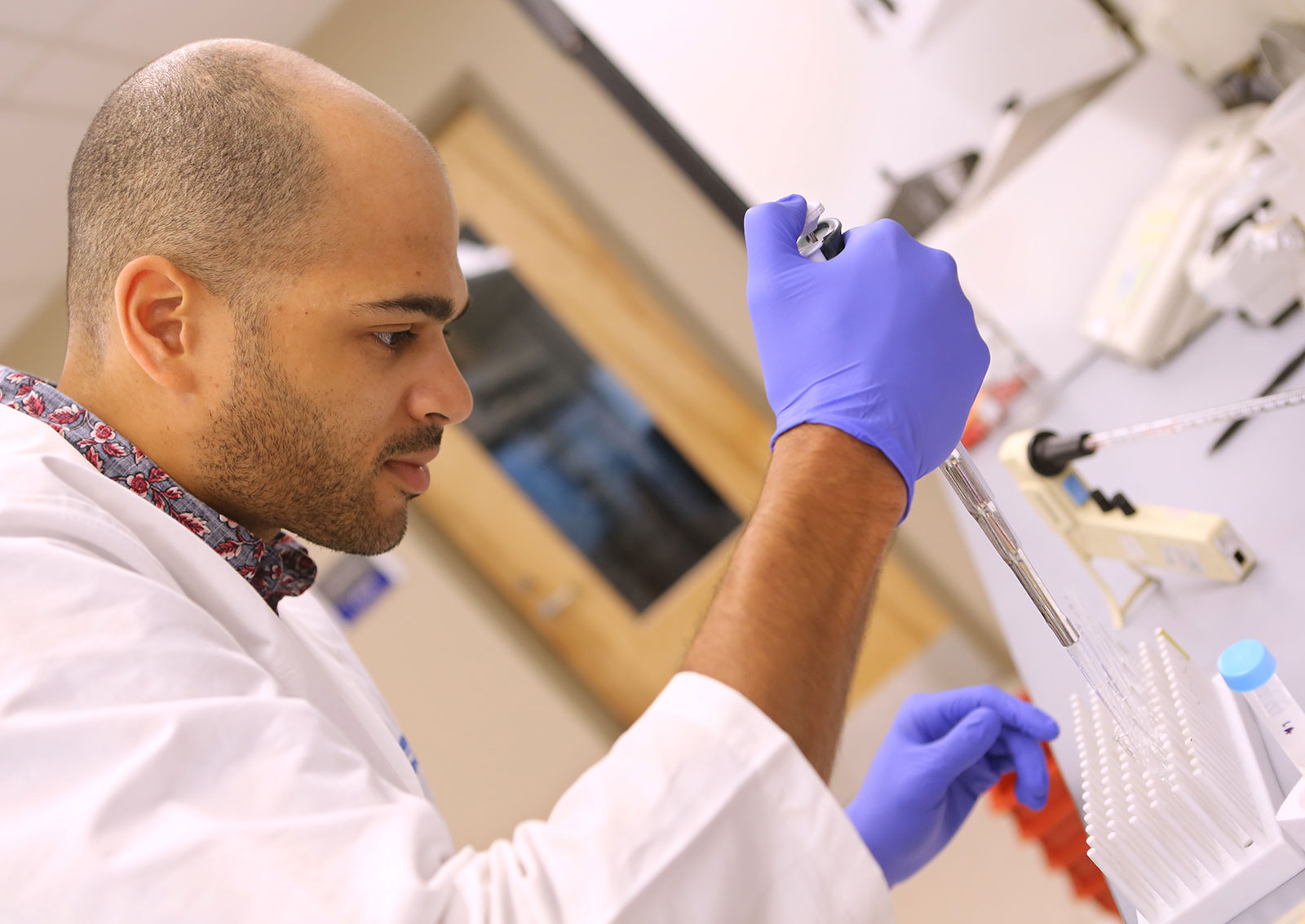 Robert Nwokonko performs research on calcium signaling in cells, which can help improve understanding of autoimmune diseases and diseases that compromise the immune system. Nwokonko is pictured wearing a laboratory coat and gloves, working at a laboratory bench with a pipette in his right hand.