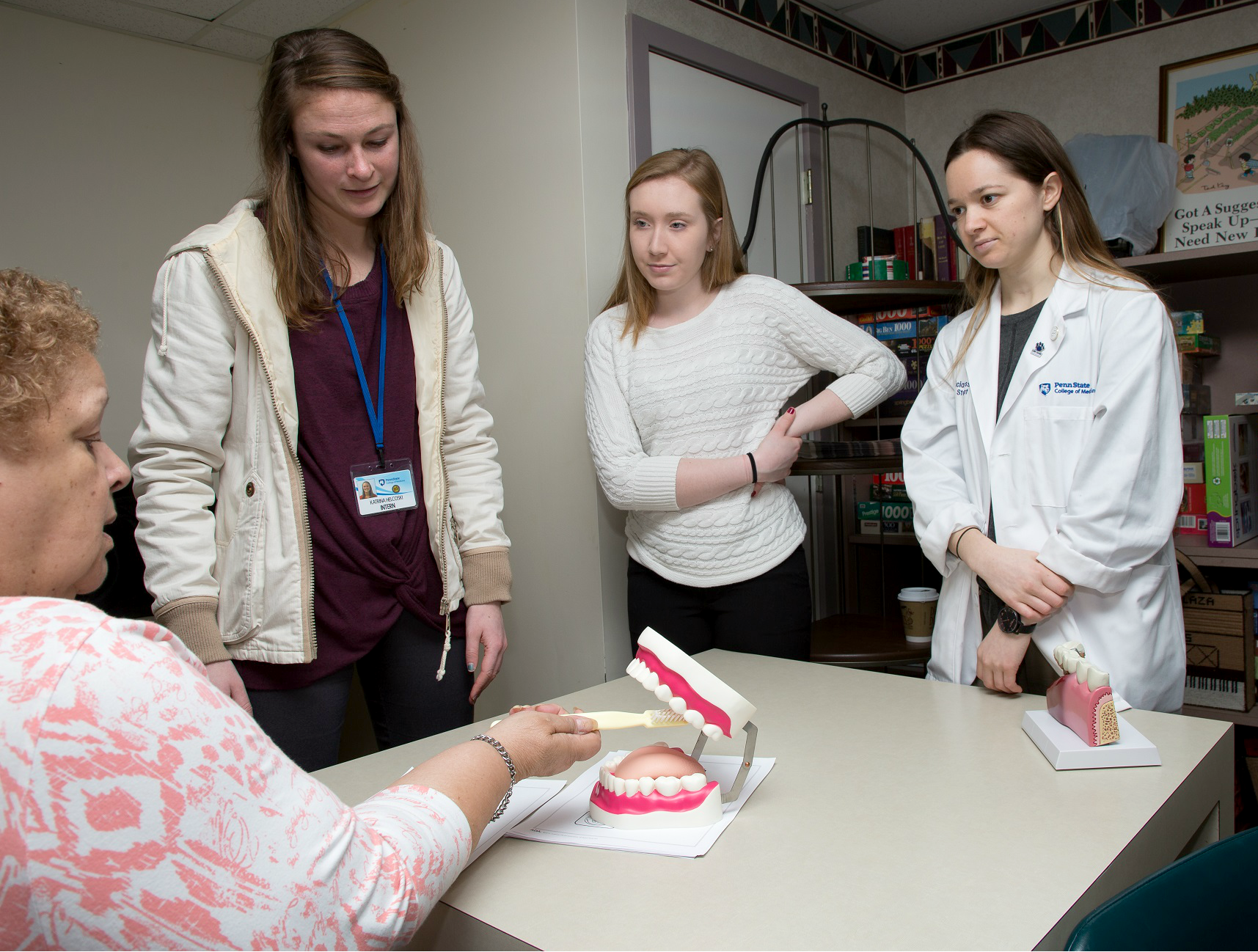 Hershey Plaza Apartments resident Millie Taylor sits at a table and practices brushing a large set of plastic teeth as Penn State College of Medicine students look on. Taylor is wearing a patterned blouse, From left, the students are wearing a sweater jacket, a sweater and a lab coat. Behind them are bookshelves filled with books and games.